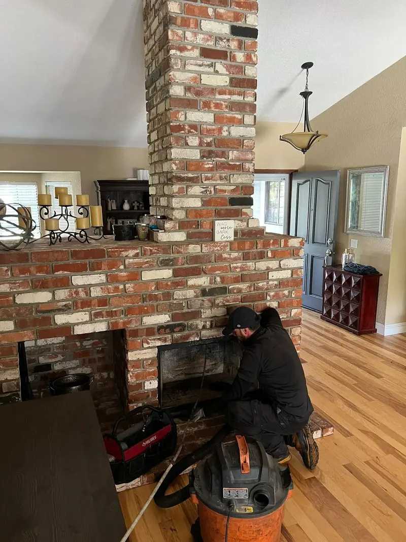 Exclusive Chimney Sweep Services technician kneeling at an open red brick fireplace in an Atlanta home, working a sweep rod into the firebox with a HEPA shop vacuum beside him